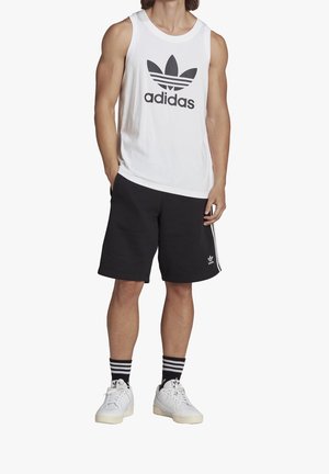 Mężczyzna ubrany w białą bezrękawnikową koszulkę Adidas, czarne spodenki Adidas w paski, czarne skarpetki w paski oraz białe tenisówki, stojący z rękami w kieszeniach.
