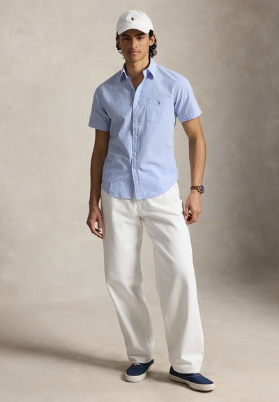 Jeune homme portant une casquette blanche, une chemise à manches courtes rayée bleue, un pantalon blanc, des chaussures bleues à enfiler et une montre-bracelet, debout devant un fond neutre.