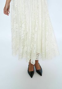 Maxikleid aus Spitze in Off-White mit aufwendigen Blumenmustern und einem fließenden Design, kombiniert mit spitzen schwarzen Lederschuhen.