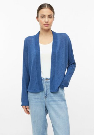 Femme portant un cardigan bleu ouvert sur un top blanc et un jean bleu clair, debout avec une main dans la poche, sur fond blanc.