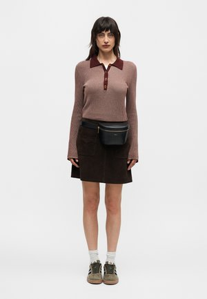 Femme portant un pull polo marron à manches longues, une jupe sombre avec des poches avant, un sac banane noir, des chaussettes blanches et des baskets beiges, debout face à l'avant.