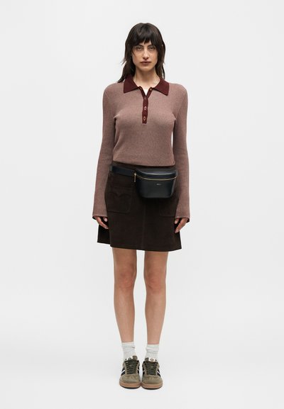 Γυναίκα που φοράει καφέ μακρυμάνικη μπλούζα πόλο, σκοτεινή φούστα με μπροστινές τσέπες, μαύρη τσαντάκι μέσης, λευκές κάλτσες και μπεζ αθλητικά παπούτσια, όρθια και κοιτάζοντας προς τα εμπρός.