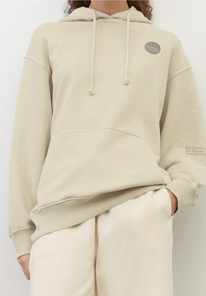 Osoba ubrana w jasnobeżową, oversize'ową bluzę z kapturem i przednią kieszenią oraz pasujące luźne spodnie, z ręką w kieszeni, na jednolitym tle.