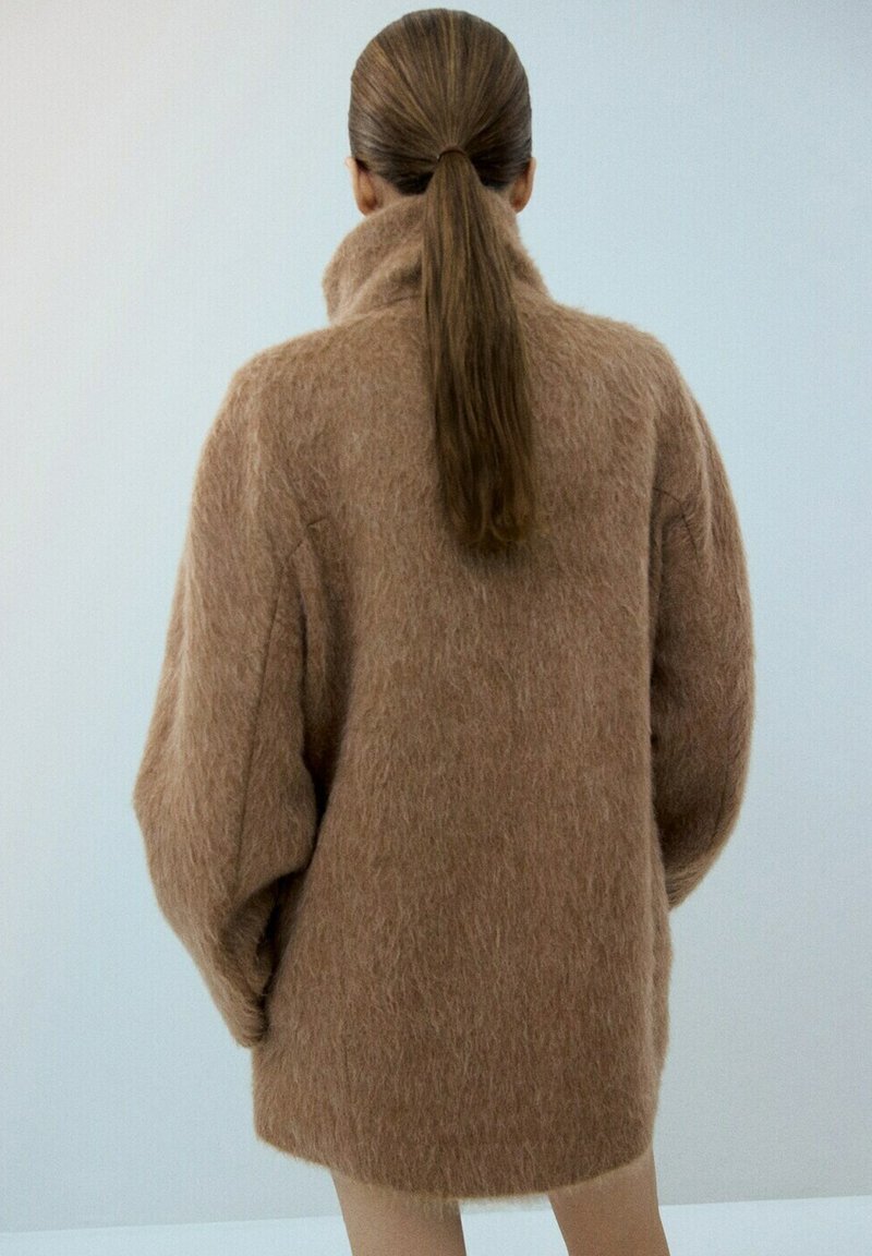 Persoană cu păr lung și brunet prins la spate, purtând o haină maro, largă și pufoasă, stând cu spatele la cameră, pe un fundal simplu, deschis la culoare.
