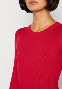 Roter gerippter Strickpullover mit Rundhalsausschnitt und langen Ärmeln, der eine glatte Textur und eine figurbetonte Silhouette aufweist.