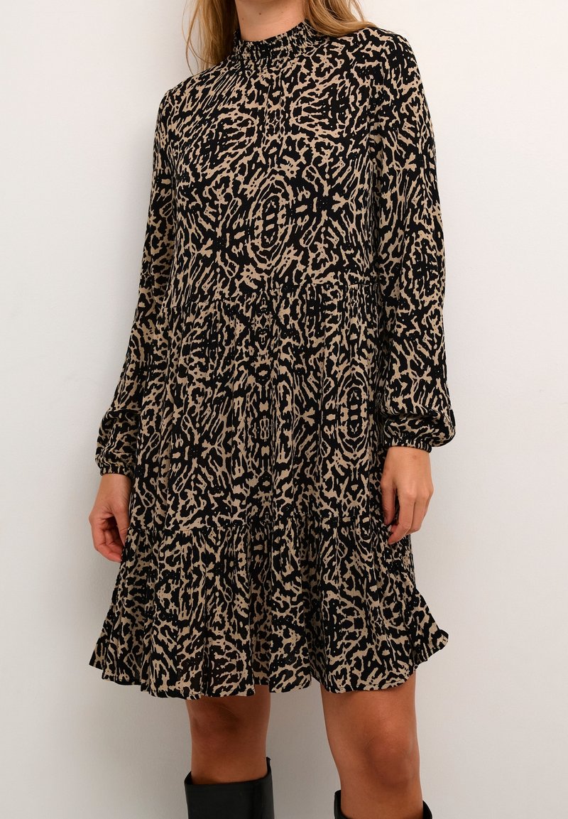 Femme portant une robe à manches longues, longueur genou, avec un motif abstrait noir et beige et un ourlet à volants, associée à des bottes noires montantes jusqu'aux genoux.