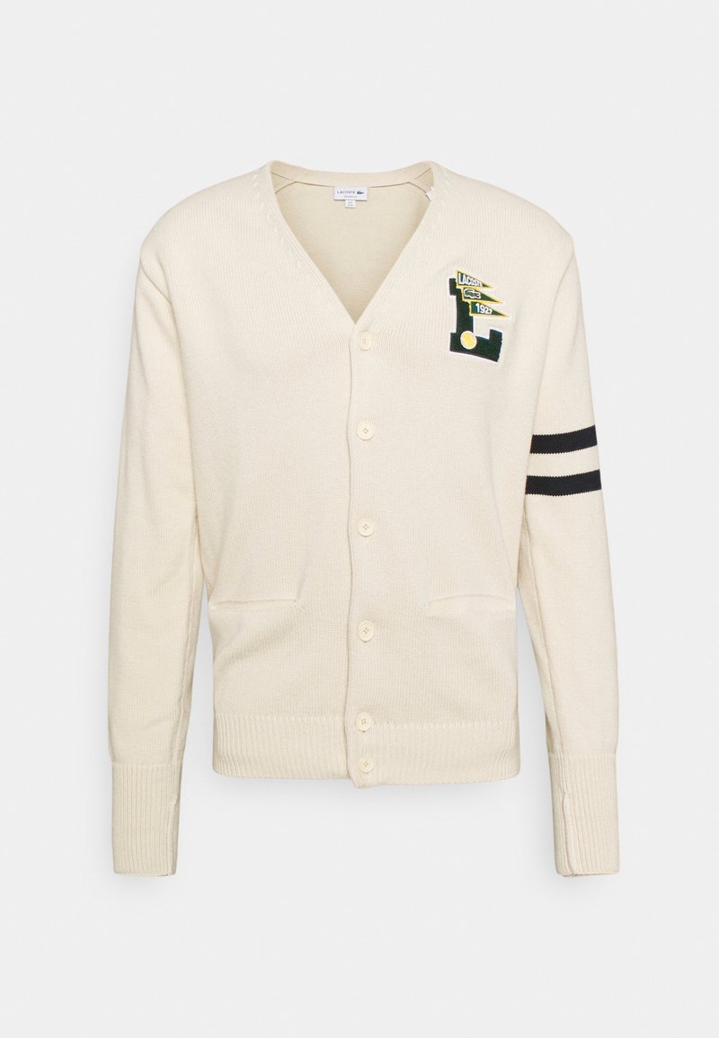 Beige cardigan made of textured knit fabric. Features a V-neck, buttons down the front, two pockets, and black stripes on the sleeves.