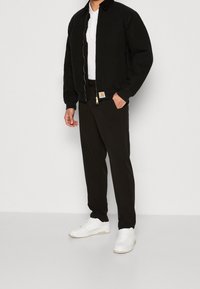 Svart bomberjacka med dragkedja, ribbad mudd och krage. Vit polo t-shirt under, svarta byxor och vita sneakers.