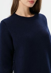 Pull en laine bleu marine avec un col rond, présentant des accents de boutons sur le côté et un tissu texturé pour plus de chaleur.