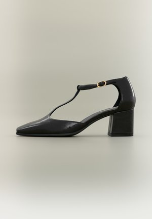 Chaussure noire en cuir verni à bride en T, talon bloc et boucle dorée sur fond neutre.