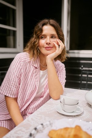 Femme en chemise rose rayée assise à une table avec des tasses à thé et un croissant, reposant son menton sur sa main et regardant sur le côté.