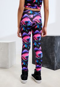Leggings neri con un motivo marmorizzato colorato e vorticoso in rosa, viola e blu. Il materiale sembra elastico; abbinati a scarpe da ginnastica nere.
