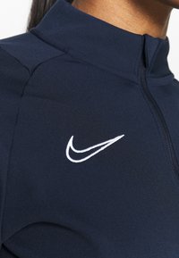 Giacca sportiva blu navy con colletto alto, chiusura a zip e logo Nike ricamato bianco sul petto sinistro. Tessuto liscio ed elasticizzato.