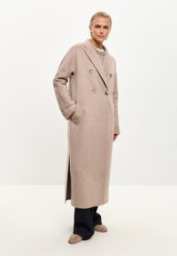 Cappotto doppiopetto beige in misto lana con grandi bottoni, tasche laterali e un design lungo e dritto. Presenta un colletto a revers e spacco laterale.