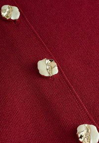 Il tessuto in maglia rossa presenta una superficie testurizzata con bottoni metallici dorati e bianchi che ricordano forme organiche. I bottoni sono di forma irregolare e lucidi.