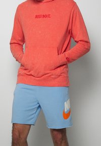 Felpa con cappuccio color corallo con scritta "JUST DO IT", abbinata a pantaloni corti azzurri chiari con un grande logo arancione Nike. Tessuto in misto cotone, vestibilità comoda.