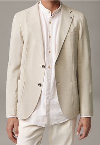 Uomo in blazer testurizzato bianco sporco sopra una camicia bianca a colletto alla mandarino con pantaloni crema, su uno sfondo neutro.