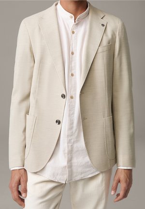 Mand med en off-white tekstureret blazer over en hvid mandarin-krave skjorte og cremefarvede bukser mod en neutral baggrund.
