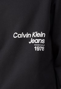 Sweat-shirt noir avec un texte blanc indiquant "Calvin Klein Jeans Established 1978." Le matériau semble doux avec une texture lisse.