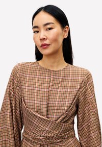 Jonge vrouw met lang zwart haar, gekleed in een bruine en roze geruite jurk met een getailleerde taille en lange pofmouwen.