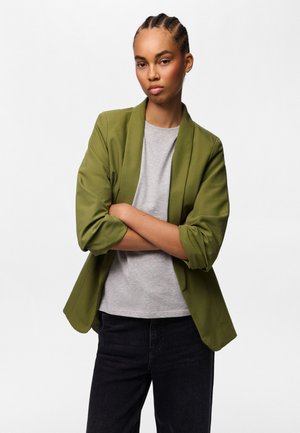 Kvinde med flettet hår iført en olivengrøn blazer over en grå skjorte og sorte bukser, stående med armene krydset mod en hvid baggrund.
