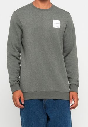Homme portant un sweat-shirt gris uni à manches longues avec un petit logo blanc Columbia sur la poitrine et un jean bleu.