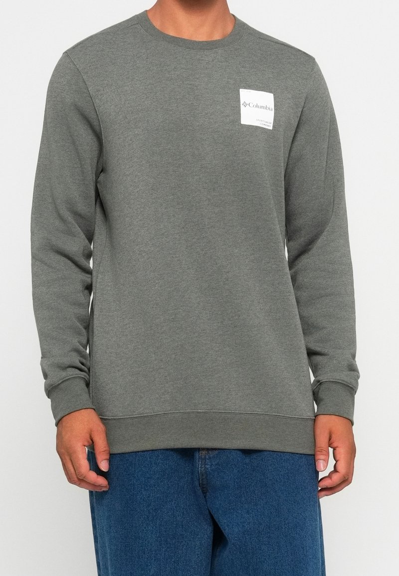 Homme portant un sweat-shirt gris uni à manches longues avec un petit logo blanc Columbia sur la poitrine et un jean bleu.