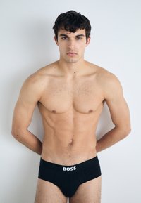 Culotte en coton noir avec logo "BOSS" en blanc. Modèle aux cheveux courts et ondulés, se tenant contre un fond neutre, les bras positionnés derrière le dos.