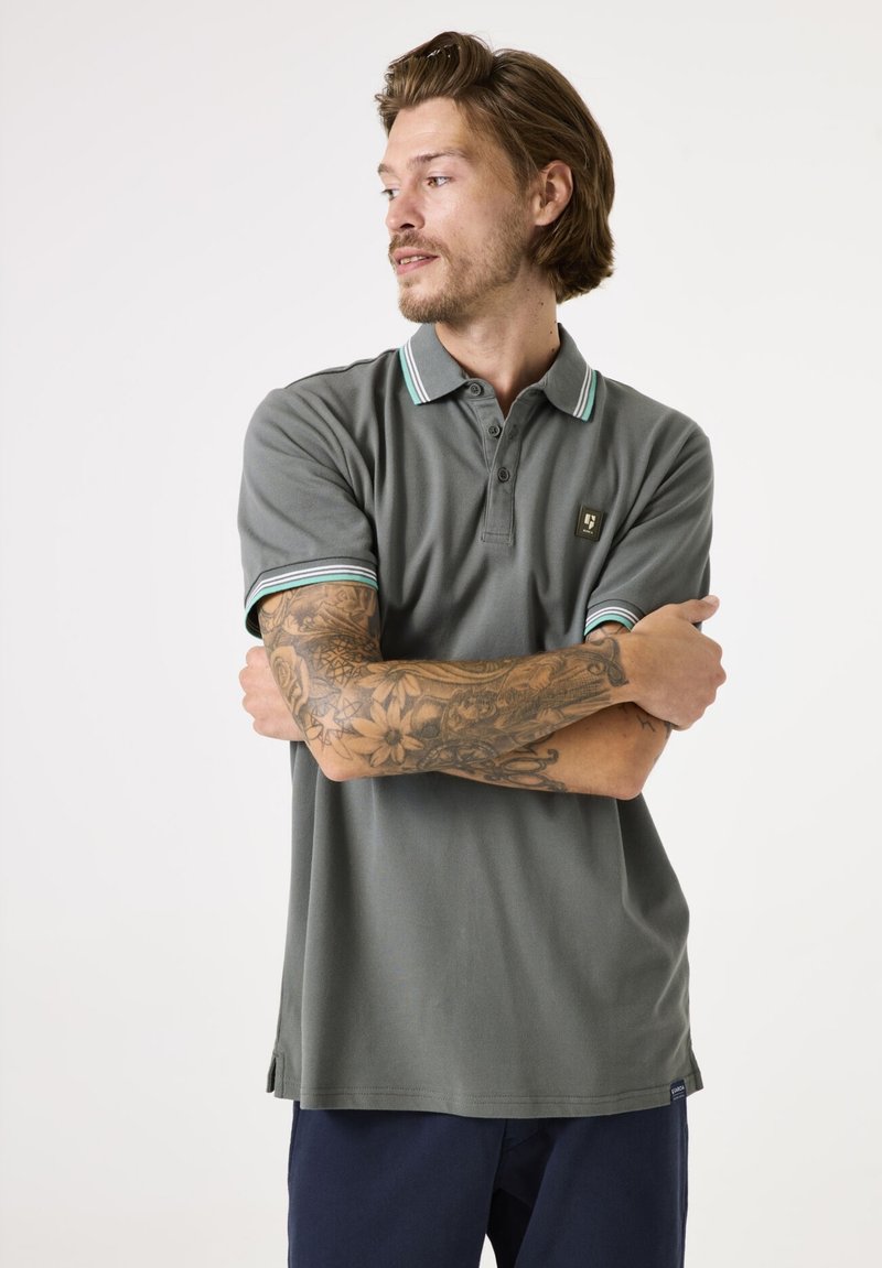 Grijze polo met een kraag met groene en witte strepen, korte mouwen en een klein logo-opslag op de linkerborst. Gladde stofstructuur.