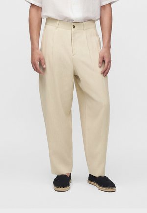 Homme portant un pantalon beige ample, une chemise blanche à manches courtes rentrée, et des espadrilles noires à enfiler sur un fond uni.