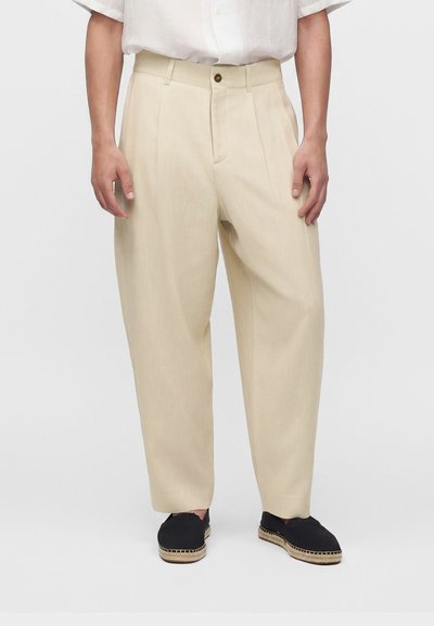Homme portant un pantalon beige ample, une chemise blanche à manches courtes rentrée, et des espadrilles noires à enfiler sur un fond uni.