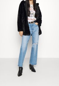 Cappotto in pelliccia sintetica nera su una maglietta bianca con grafica, abbinato a jeans dritti azzurri chiari con dettagli strappati e stivali neri alla caviglia.