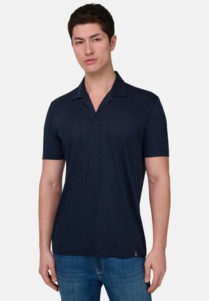 Mand iført en tætsiddende marineblå kortærmet polo og blå jeans, stående med afslappede arme mod en ensfarvet lys baggrund.