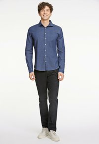 Camicia button-up da uomo di colore blu scuro, con colletto, maniche lunghe e bottoni beige, abbinata a pantaloni neri e sneaker chiare.