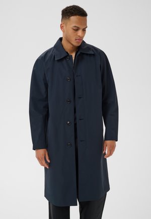 Mann trägt einen langen, dunkelblauen, geknöpften Mantel mit Kragen, steht vor einem einfarbigen hellen Hintergrund und schaut nach unten.