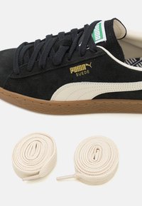 Czarny zamszowy sneakers z białymi akcentami, brązową gumową podeszwą, złotym logo i czarnymi sznurówkami. Zawiera dwie zwinięte kremowe sznurówki.