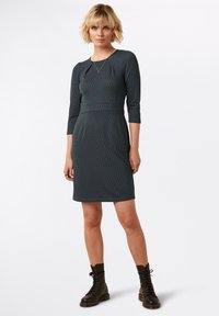 Laivastonsininen mekko, jossa on geometrinen kuvio, kolmen neljänneksen hihat, tiukka vyötärö ja polvenpituinen helma. Tämä yhdistetään mustiin nauhallisiin saappaisiin.