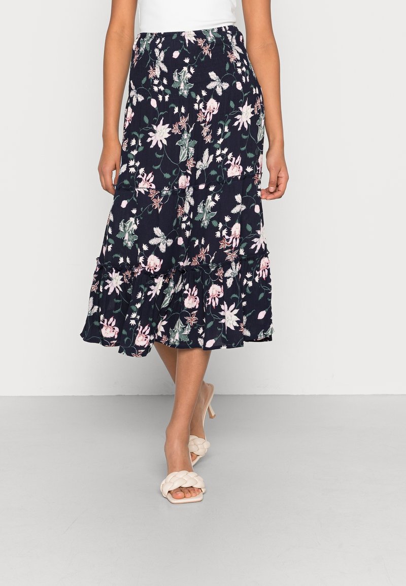 Femme portant une jupe midi bleu marine à motif floral blanc et rose, associée à des sandales blanches à talons tressées et un haut blanc.