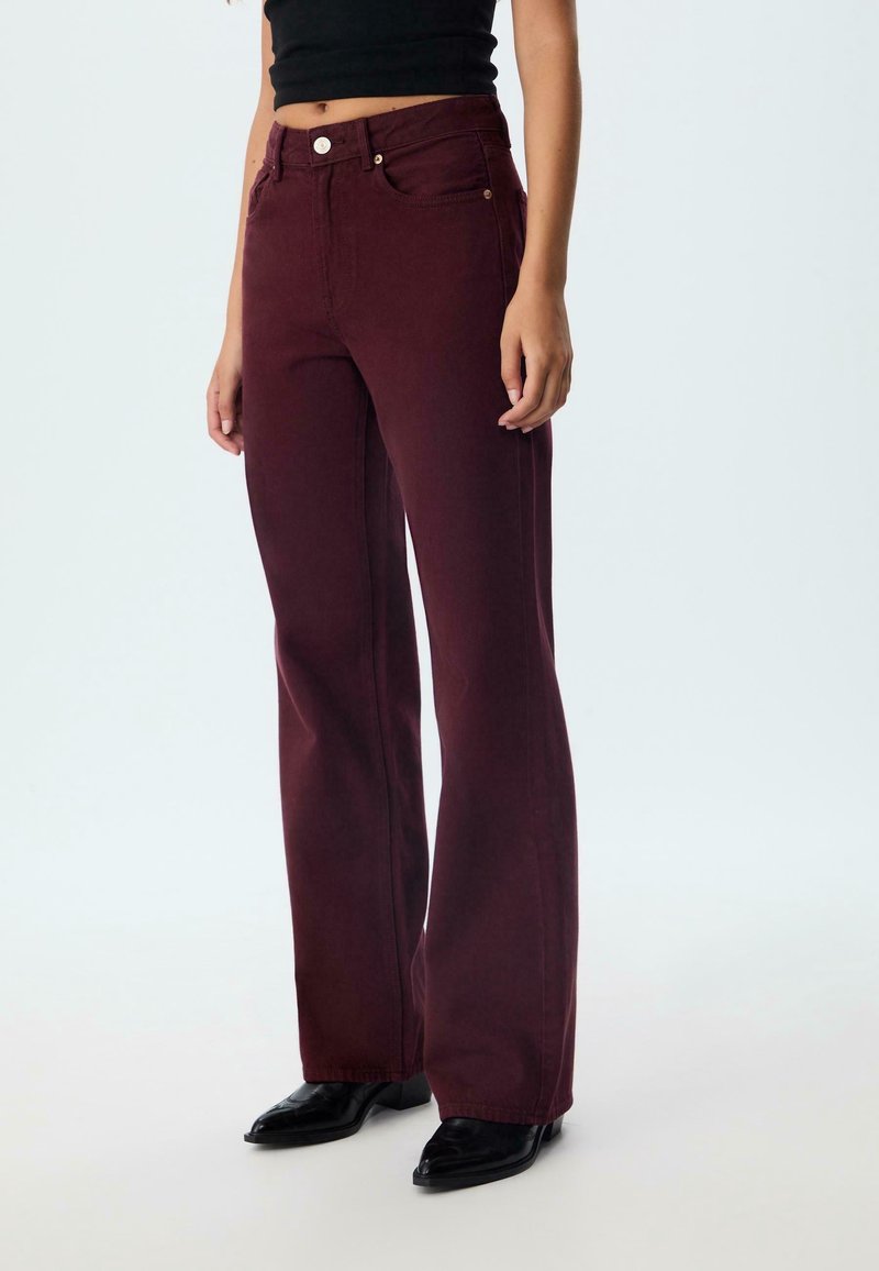 Femme portant un pantalon bordeaux taille haute à jambes droites, des chaussures noires pointues et un haut court noir, debout devant un fond uni.