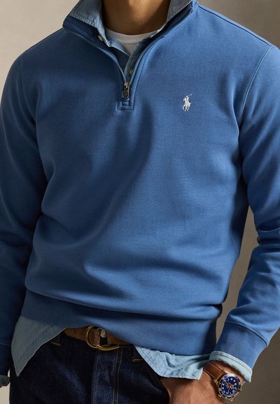 Μπλε πουλόβερ με τεταρτημόριο φερμουάρ, κατασκευασμένο από απαλό ύφασμα. Διαθέτει λευκό λογότυπο, φορεμένο πάνω από ανοιχτό γαλάζιο πουκάμισο με γιακά.