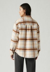 Fleece plaid-paita vaaleancremessä, ruskeassa ja sinisessä, rentossa leikkauksessa, napitettuissa hihoissa ja kaarevassa helmassa. Sisältää kontrastivärisen ruutukuvion.