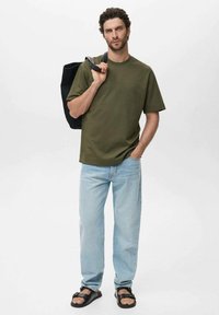 Olive green cotton t-shirt, relaxed fit, paired with light blue denim jeans. Black tote bag and black sandals accent the outfit.