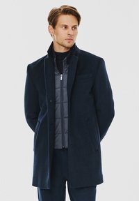 Granatowy płaszcz z wełny z domieszką, z klapami, z pojedynczym guzikiem, dopasowany krój oraz kieszenie boczne, noszony na szarym pikowanym kurtce.