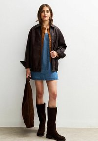 Mujer vestida con una chaqueta de cuero marrón, un cárdigan naranja abotonado, un vestido de mezclilla azul y botas altas de cuero marrón oscuro hasta la rodilla, sosteniendo un bolso marrón a juego.