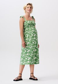 Grön blommig klänning med en smockad midja, tunna axelband med rosetter och en knälång A-linjeformad kjol. Svarta platta sandaler fulländar looken.