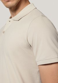 Beżowa koszulka polo z teksturowanej tkaniny, z dwu-przyciskowym zapięciem i krótkimi rękawami. Kołnierz i mankiety są starannie wykończone.