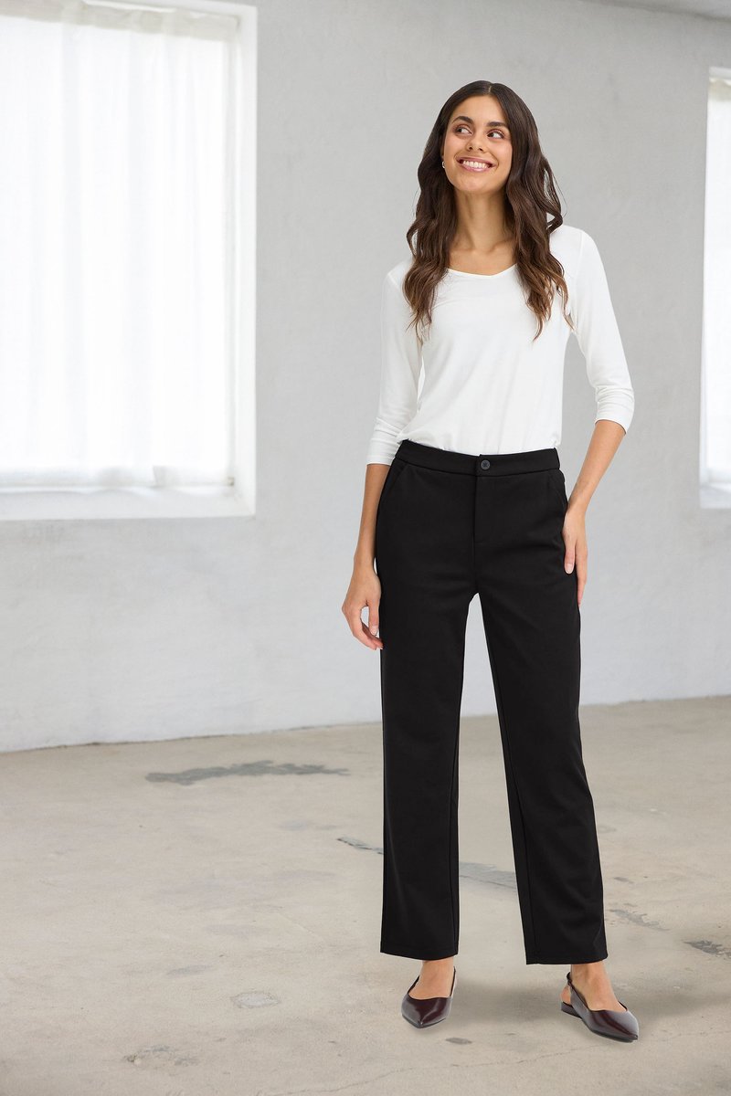 Woman standing indoors wearing white long-sleeve top, black pants, and dark pointed shoes, smiling and looking to her left.