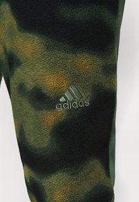 Fleece-housut, joissa on vihreä, musta ja ruskea maastokuvio. Vasemmassa reidessä on brodeerattu Adidas-logo.