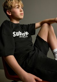 Sort t-shirt med hvid "Subserie LDN 51 6289" tekst, parret med sorte shorts, siddende stilling, kortærmede og hvide stribede sokker.