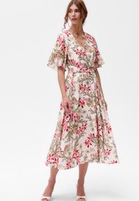 Frau, die ein weißes Midi-Kleid mit pinken und grünen Blumenmuster, schwingenden Ärmel, V-Ausschnitt und Taillengürtel präsentiert, kombiniert mit weißen High Heels.